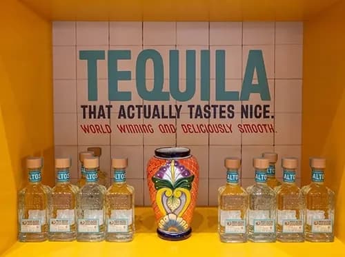 Altos bottles arranged on a yellow shelf against a tiled TEQUILA backdrop panel